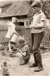 Vaikaičiai Omos Marės Reizgienės gėlių darželyje, kuris aptvertas tvorele. Sesutė Ina laisto gėles, jauniausias broliukas Gintaras tupi su žąsiuku rankose, vyriausias Albertas lesina žąsiukus. Už tvorelės matosi staliaus kamara, kurioje Opa mėgo meistrauti, anksčiau buvo pagaminęs baldus. Apie 1970 m. Fotografas nežinomas / Iš I. E. Kuzmienės šeimos albumo